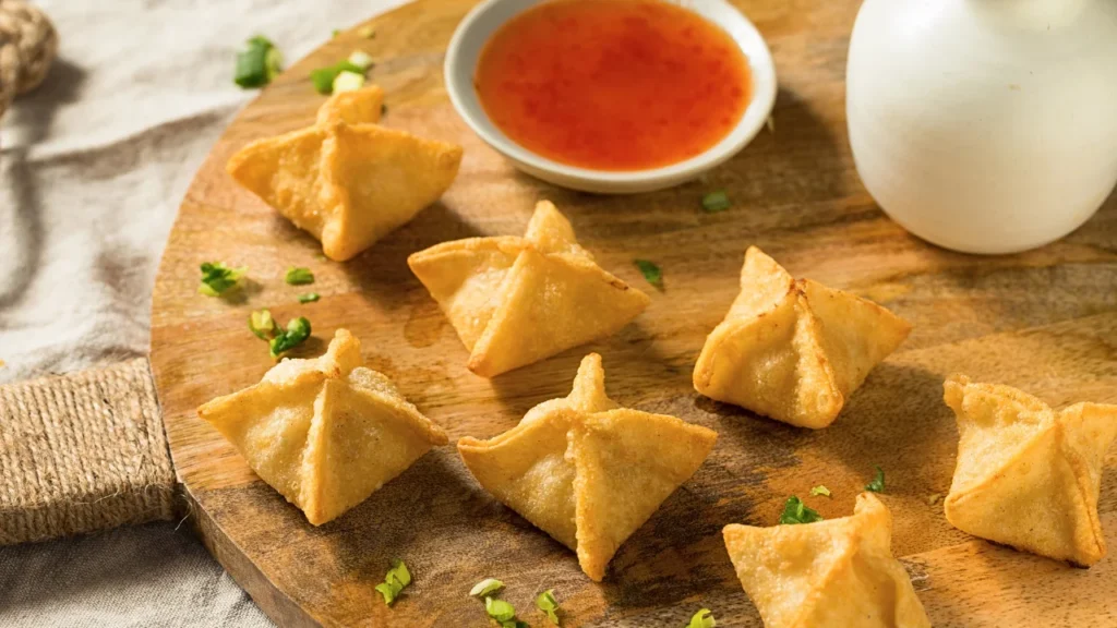 Golden brown Crab Rangoon arranged on a white plate with a small bowl of sweet and sour dipping sauce.
