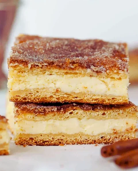 A stack of churro cheesecake bars with a creamy layer between crispy churro crusts, in a light, simple setting.